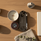 A candle with a pink container and a silver 3-piece accessory set placed on a tray, all presented on a neutral background.