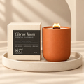 A lit orange terracotta candle in a matte ceramic holder sits beside its cream-colored packaging box with a black label reading 'Citrus Kush' and 'KQO Pure Organics,' placed on a round beige tray against a warm beige background with soft shadows.